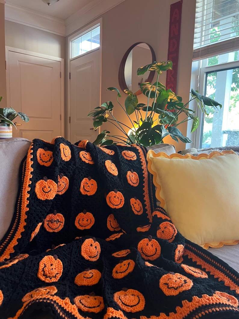May include: A black and orange crocheted blanket featuring smiling pumpkin faces. The blanket has an orange border and is draped over a sofa. A yellow pillow with a scalloped edge is also on the sofa. The blanket is a festive Halloween decoration.