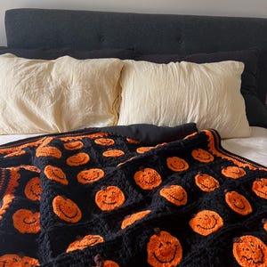 May include: A black crochet blanket featuring smiling orange pumpkins. The blanket is draped over a bed with cream-colored pillows and a dark gray headboard. The pumpkins have brown stems and black smiles.