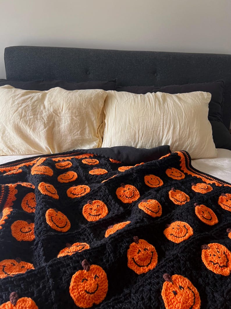 May include: A black crochet blanket featuring smiling orange pumpkins. The blanket is draped over a bed with cream-colored pillows and a dark gray headboard. The pumpkins have brown stems and black smiles.