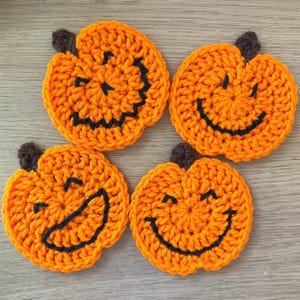 May include: Four handmade, orange crochet pumpkin coasters with black stitched faces and brown stems. Each coaster features a different jack-o'-lantern face design. These festive coasters are perfect for Halloween or autumn decor.