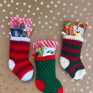 Puede incluir: Tres calcetines navideños hechos a mano en rojo, verde y blanco, llenos de pequeños regalos y bastones de caramelo. Los calcetines están tejidos a ganchillo con un punto texturizado y tienen puños y talones blancos. El fondo es dorado con motivos de copos de nieve.