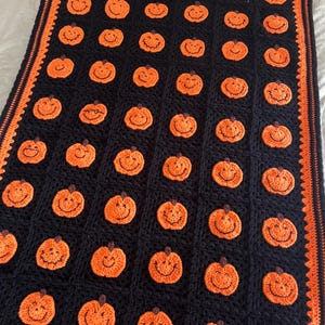 May include: A handmade crochet blanket featuring a repeating pattern of smiling orange pumpkins on a black background. The blanket has an orange border and is perfect for Halloween or autumn decor. The blanket is made of soft yarn.