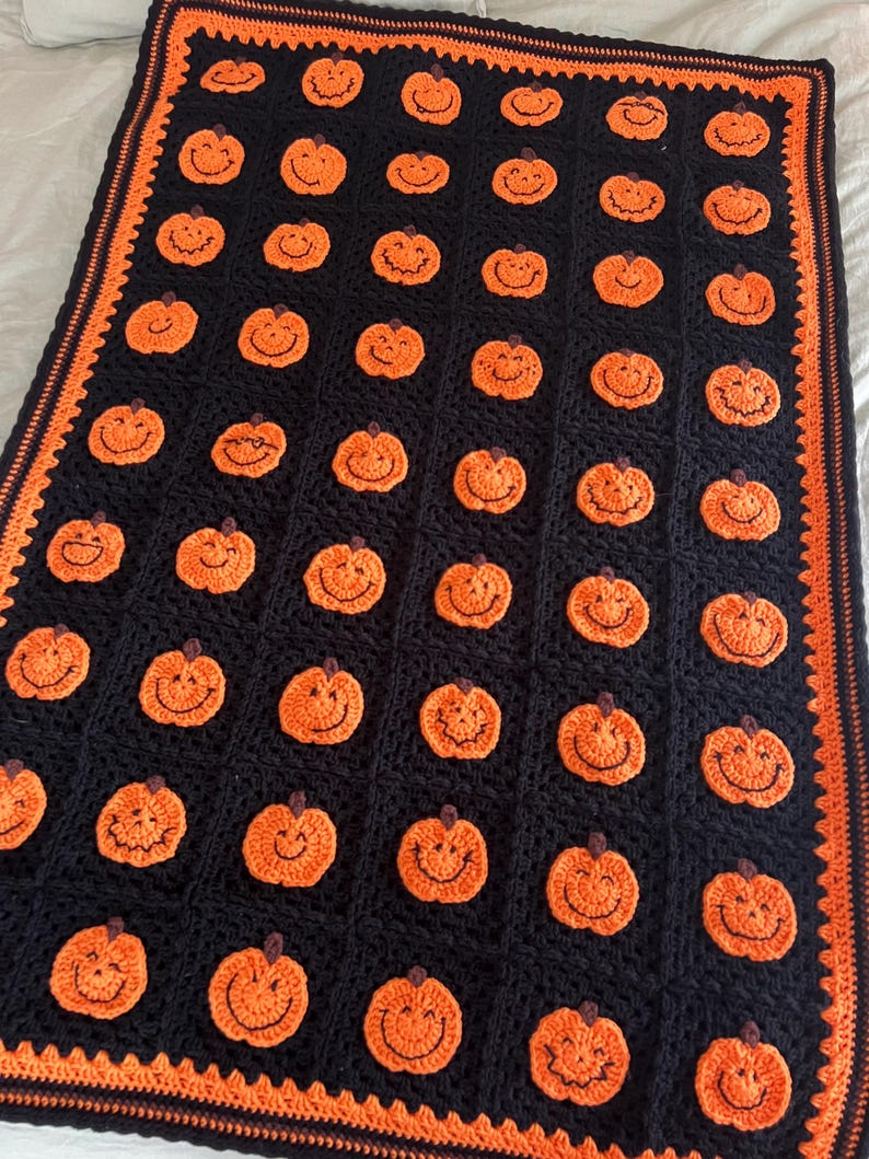 May include: A handmade crochet blanket featuring a repeating pattern of smiling orange pumpkins on a black background. The blanket has an orange border and is perfect for Halloween or autumn decor. The blanket is made of soft yarn.