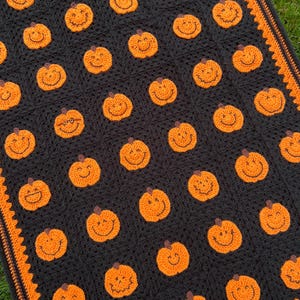 May include: A black crochet blanket featuring numerous orange pumpkin faces with smiling expressions. The blanket has an orange border and is likely handmade, perfect for Halloween or autumn decor.