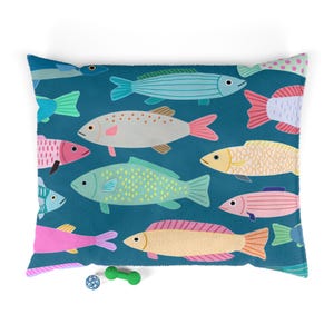 May include: A square throw pillow with a deep teal background featuring a repeating pattern of stylized fish in various colors including light blue, yellow, pink, and white. The fish have distinct patterns like dots and stripes.