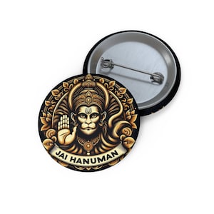 May include: A gold and black button featuring a detailed illustration of the Hindu deity Hanuman. The image shows Hanuman with his hand raised in a gesture of blessing. The text "Jai Hanuman" is written in a banner below the image.
