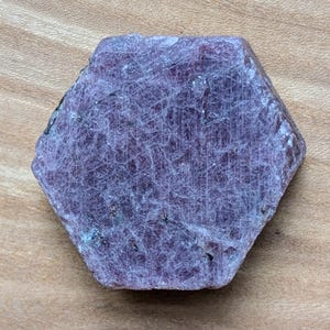 May include: A hexagonal, raw ruby gemstone with a deep red-purple hue. The surface displays a network of fine lines and subtle variations in color. The stone is set against a light brown wooden surface.
