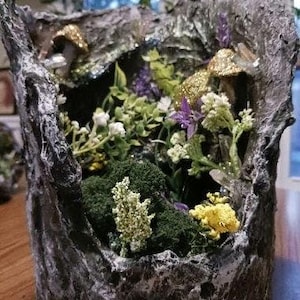 May include: A miniature fairy garden scene inside a gray, textured, tree trunk-shaped planter. The scene includes a small, mossy forest with purple and white flowers, and gold-colored mushrooms.
