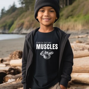 May include: A young person wearing a black t-shirt with the text "CHECK OUT MY MUSCLES" and a graphic of three mussels. The person is also wearing a black beanie and brown pants.