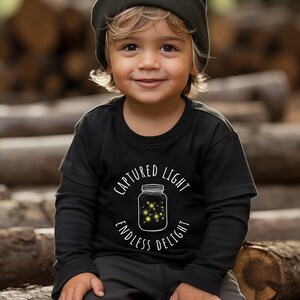 Pode incluir: Uma criança pequena vestindo uma camisa preta de manga comprida com um gráfico branco de um frasco cheio de vaga-lumes. O texto na camisa diz "Captured Light Endless Delight". A criança está usando um gorro preto e está sentada em uma pilha de toras.