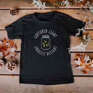 Op de afbeelding: Zwart T-shirt met een witte afbeelding van een pot met vuurvliegjes erin. De tekst rond de pot luidt "Captured Light Endless Delight".