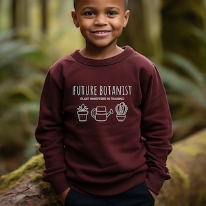 Könnte beinhalten: Ein maroonfarbener Pullover mit dem Text "Future Botanist Plant Whisperer in Training" in Weiß. Das Design beinhaltet eine Topfpflanze, eine Gießkanne und einen Kaktus.