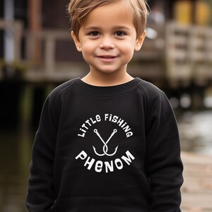 May include: A black sweatshirt with the text "Little Fishing Phenom" in white. The design includes two crossed fishing hooks. The sweatshirt is worn by a young child.