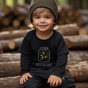 Op de afbeelding: Een zwart longsleeve shirt met een witte afbeelding van een pot vol met vuurvliegjes. De tekst "Catch the Light" is onder de pot gedrukt. Het shirt wordt gedragen door een kind met een groene beanie.