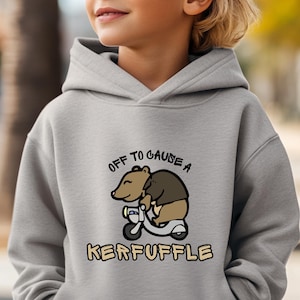 May include: A light gray hooded sweatshirt featuring a cartoon bear riding a scooter. The text "OFF TO CAUSE A KERFUFFLE" is printed above and below the bear illustration in a playful font. The sweatshirt is made of a soft material.