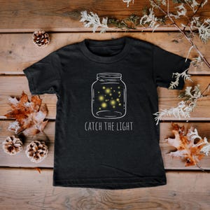 Op de afbeelding: Zwart T-shirt met een witte omtrek van een glazen pot gevuld met vuurvliegjes en de tekst "CATCH THE LIGHT".