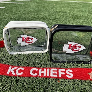 May include: Two clear stadium-approved bags, one white and one black, each with a Kansas City Chiefs logo. A red strap with "KC CHIEFS" in white lettering lies below the bags on a green field.