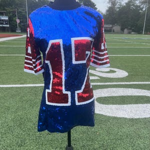 May include: A sequined jersey-style top in blue, red, and white, displayed on a mannequin. The number "17" is prominently featured on the front. The sleeves have white stripes. The top is on a black stand, set against a green field.
