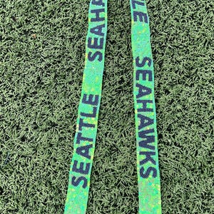 May include: A green beaded strap with the words "SEATTLE SEAHAWKS" in black lettering. The strap has silver-colored metal clasps at the ends. The background is green.