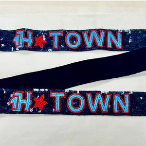 May include: A dark blue sequined bag strap featuring the text H-TOWN in light blue beaded letters with a red star between the H and TOWN. The strap includes gold-toned metal lobster claw clasps at both ends for secure attachment to a handbag.