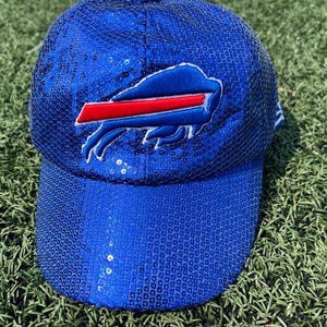 May include: A blue baseball cap covered in sequins, featuring the Buffalo Bills logo in white, red, and blue. The cap is on a green grassy surface.