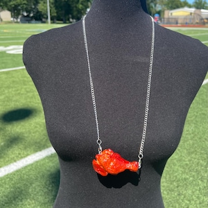 May include: A silver chain necklace featuring a vibrant, red-orange chicken drumstick pendant. The pendant is detailed with realistic textures and coloring, creating a unique and eye-catching accessory. The necklace is displayed on a black mannequin.