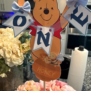 Op de afbeelding: Een Winnie de Poeh-thema taarttopper. Het personage houdt banners vast met de tekst "ONE". De topper bevat een honingpot met een bij en is omringd door roze en perzikrozen.
