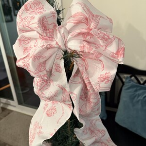 May include: A large, decorative bow made of white fabric with a pink floral pattern, attached to a Christmas tree. The bow is voluminous and features a detailed botanical design, adding a touch of elegance to holiday decor.