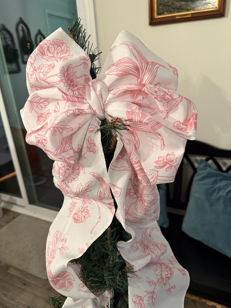 May include: A large, decorative bow made of white fabric with a pink floral pattern, attached to a Christmas tree. The bow is voluminous and features a detailed botanical design, adding a touch of elegance to holiday decor.