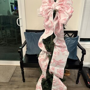 May include: A small Christmas tree decorated with a large, white and pink floral patterned bow. The bow cascades down the tree, adding a decorative touch. The tree is in a copper-colored pot, and the background includes a black bench and blue pillows.