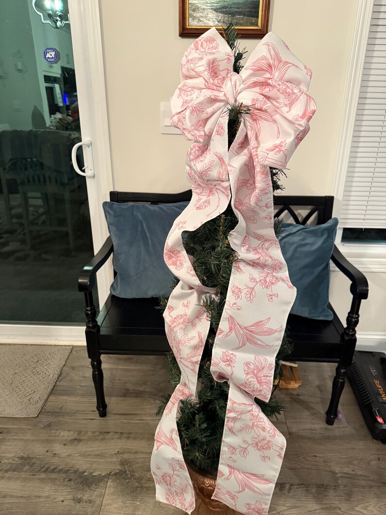 May include: A small Christmas tree decorated with a large, white and pink floral patterned bow. The bow cascades down the tree, adding a decorative touch. The tree is in a copper-colored pot, and the background includes a black bench and blue pillows.