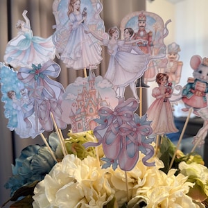 Può includere: Una composizione floreale con ritagli decorativi di ballerine, un castello e scarpette da ballo. I ritagli sono in colori pastello e attaccati a bastoncini di legno. I fiori sono bianchi, blu e verdi.