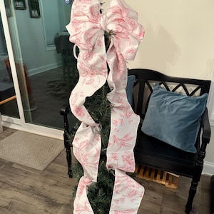 May include: A small Christmas tree decorated with a large bow made of white fabric with a pink floral pattern. The bow's long, flowing ribbons cascade down the tree. The tree is in a decorative pot, and a black bench with a blue pillow is in the background.