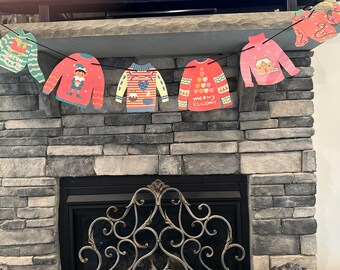 Pode incluir: Uma guirlanda de Natal com seis enfeites de suéter vermelho e verde. Os suéteres têm designs diferentes, incluindo um boneco de neve, uma árvore de Natal e um duende. A guirlanda está pendurada acima de uma lareira.