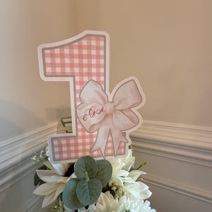 Op de afbeelding: Roze en witte ruitjes nummer één met een witte strik en de naam "E.G." erop. Het nummer één staat op een wit bloemstuk.