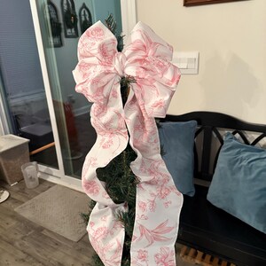 May include: A decorative Christmas tree bow made of white fabric with a pink floral pattern. The bow is large and has long, flowing tails. The bow is attached to a small, artificial Christmas tree.