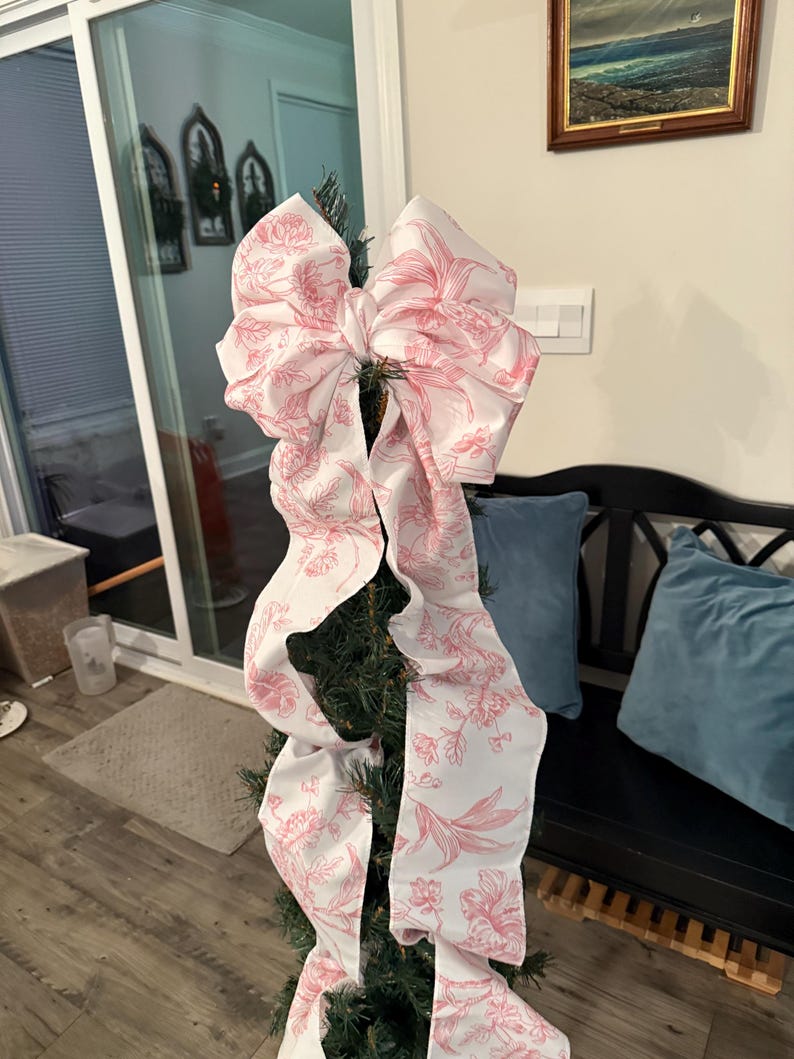 May include: A decorative Christmas tree bow made of white fabric with a pink floral pattern. The bow is large and has long, flowing tails. The bow is attached to a small, artificial Christmas tree.