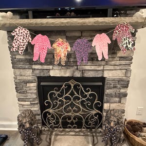 Puede incluir: Una guirnalda de seis peleles para bebés con varios estampados de animales, incluyendo leopardo y cebra, cuelga sobre una chimenea. Los peleles son en tonos rosa, morado y beige. La chimenea es de piedra gris.