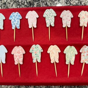May include: Twelve cupcake toppers featuring a baby onesie design. The onesies are printed with various floral patterns in pink, blue, green, and white. The toppers are attached to wooden sticks.