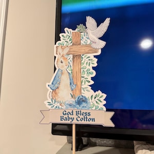 Può includere: Una croce decorativa in legno con un coniglietto, una colomba e elementi floreali. Lo striscione recita "God Bless Baby Colton". Il design presenta illustrazioni in stile acquerello in morbidi toni blu e verdi.