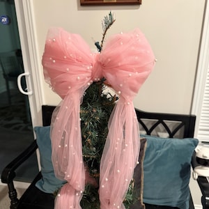 May include: A large pink tulle bow with faux pearl embellishments, used as a Christmas tree topper. The bow is tied around the top of a small evergreen tree. The bow's long streamers fall down the tree, creating a festive holiday decoration.