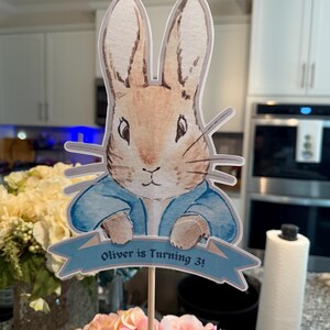 Op de afbeelding: Een taarttopper met Peter Rabbit, geïllustreerd in aquarel, met een blauw jasje. Een blauwe banner hieronder leest "Oliver is Turning 3!". De topper staat op een bed van roze en perzik bloemen.