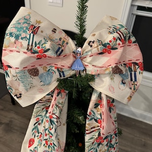 May include: A white fabric Christmas tree bow with a pink and white striped ribbon, a red stitched seam, and a festive print of a nutcracker, a Christmas tree, and other holiday imagery. A small blue and white figurine of a ballerina is attached to the bow.