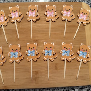 Op de afbeelding: Elf katvormige cupcake-toppers op houten spiesjes. De toppers zijn bruin met witte accenten, met roze of blauwe strikjes. Ze zijn gerangschikt op een houten snijplank.