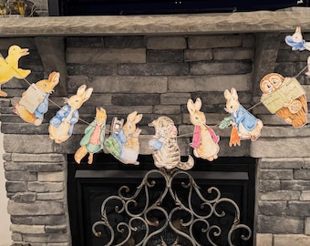 May include: A Peter Rabbit themed party banner with characters from the book, including Peter Rabbit, Benjamin Bunny, and Mrs. Tiggy-Winkle. The banner is strung across a brick fireplace.