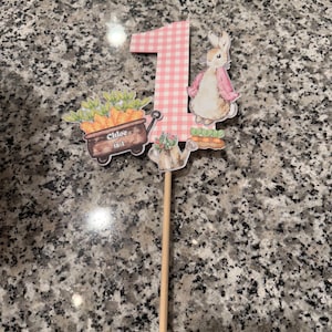 May include: A decorative cake topper featuring a pink gingham number 1, a bunny in a pink jacket, and a cart of carrots. The cart has the text "Chloe is 1". The topper is attached to a wooden dowel.