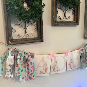 May include: A pink and white banner with the name "JADE" spelled out in large letters. Each letter has a small illustration of a bunny rabbit. The banner is strung with fabric scraps in various patterns and colors.