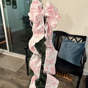 May include: A small Christmas tree decorated with a large white and pink floral patterned bow. The bow's long ribbons cascade down the tree. The tree is in a decorative pot, and the background includes a black bench and a framed picture.