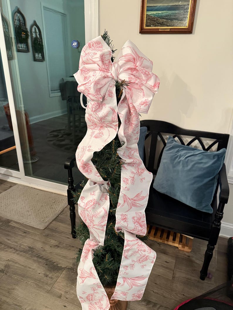 May include: A small Christmas tree decorated with a large white and pink floral patterned bow. The bow's long ribbons cascade down the tree. The tree is in a decorative pot, and the background includes a black bench and a framed picture.