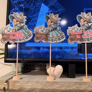 May include: Three Easter bunny decorations. Each bunny is wearing a blue dress and hat, and pushing a cart filled with Easter eggs. The text "Happy Easter" and "Some bunny is One!" are displayed on the decorations. The decorations are on wooden sticks.